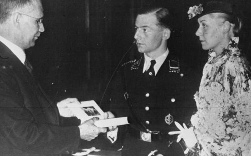 A wedding ceremony in Germany in 1936. Translation of caption: "Chancellor Hitler has decided that a copy of his book Mein Kampf will be given to every couple on their wedding day.", 1936, Bibliothèque nationale de France, Wikimedia Commons, Public Domain. 