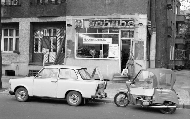 Straßenszene, im Vordergrund u.a. ein Trabant, im Hintergrund ein Schuhgeschäft