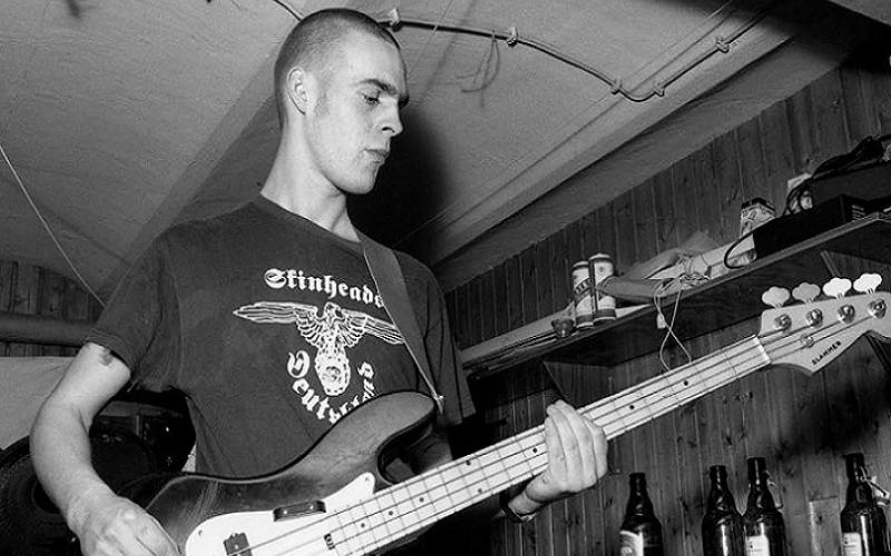 Mitglied einer Naziband spielt Gitarre und trägt ein T-Shirt mit Reichsadler und Aufschrift "Skinheads Deutschland", schwarzweiß