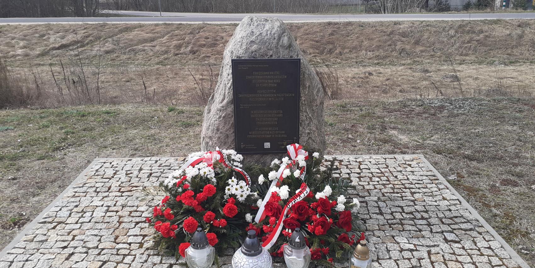 Denkmal an dem Ort der ehemaligen Bahnhofsstation im Dorf Treblinka, 31. März 2026. Die Kränze wurden vom Pilecki Institut (Witold Pilecki Institute of Solidarity and Courage) und vom Bürgermeister und den Bewohnern der Gemeinde Małkinia gestiftet; rechts eine goldene Kerze mit der goldenen Inschrift „Treblinka Museum – wir erinnern” 