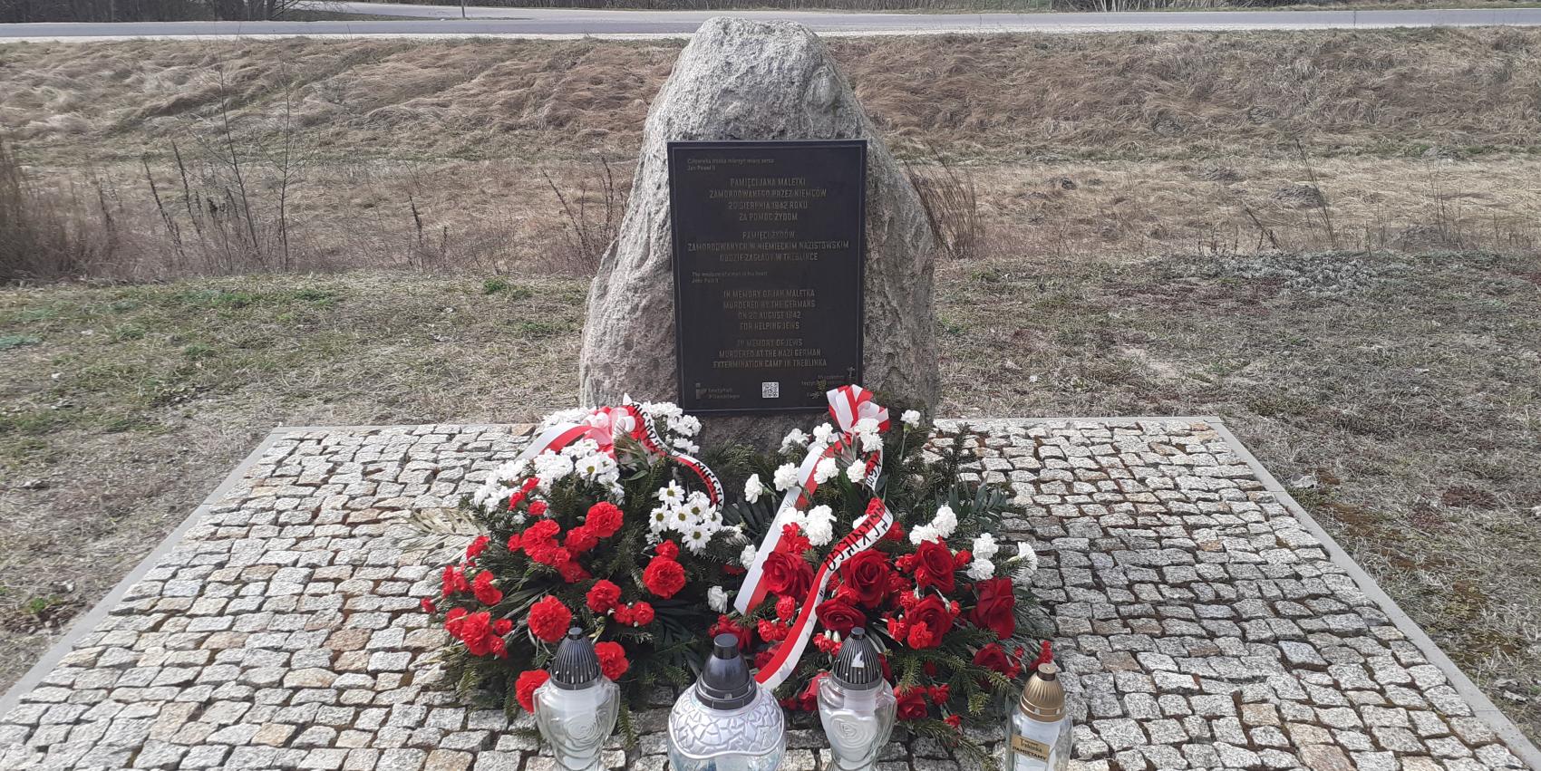 Denkmal an dem Ort der ehemaligen Bahnhofsstation im Dorf Treblinka, 31. März 2026. Die Kränze wurden vom Pilecki Institut (Witold Pilecki Institute of Solidarity and Courage) und vom Bürgermeister und den Bewohnern der Gemeinde Małkinia gestiftet; rechts eine goldene Kerze mit der goldenen Inschrift „Treblinka Museum – wir erinnern” 
