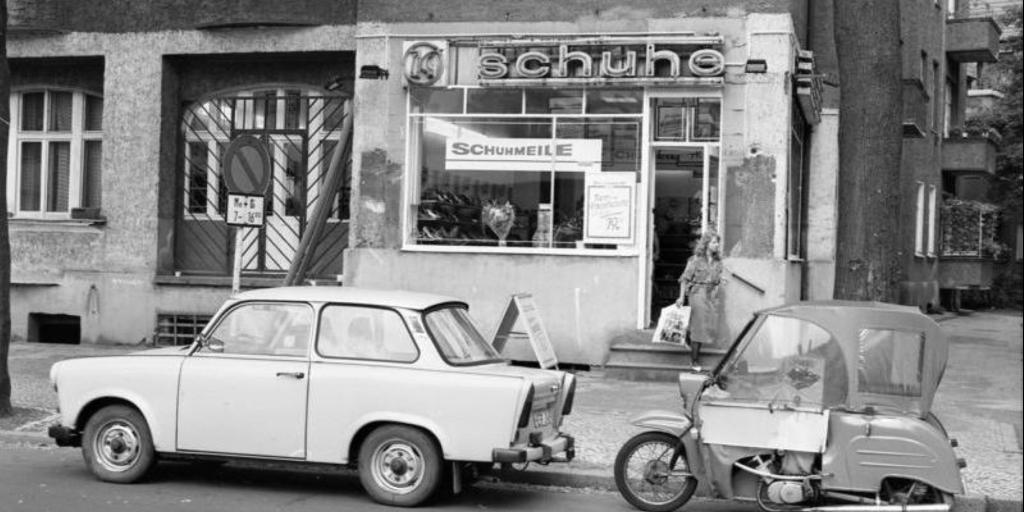 Straßenszene, im Vordergrund u.a. ein Trabant, im Hintergrund ein Schuhgeschäft