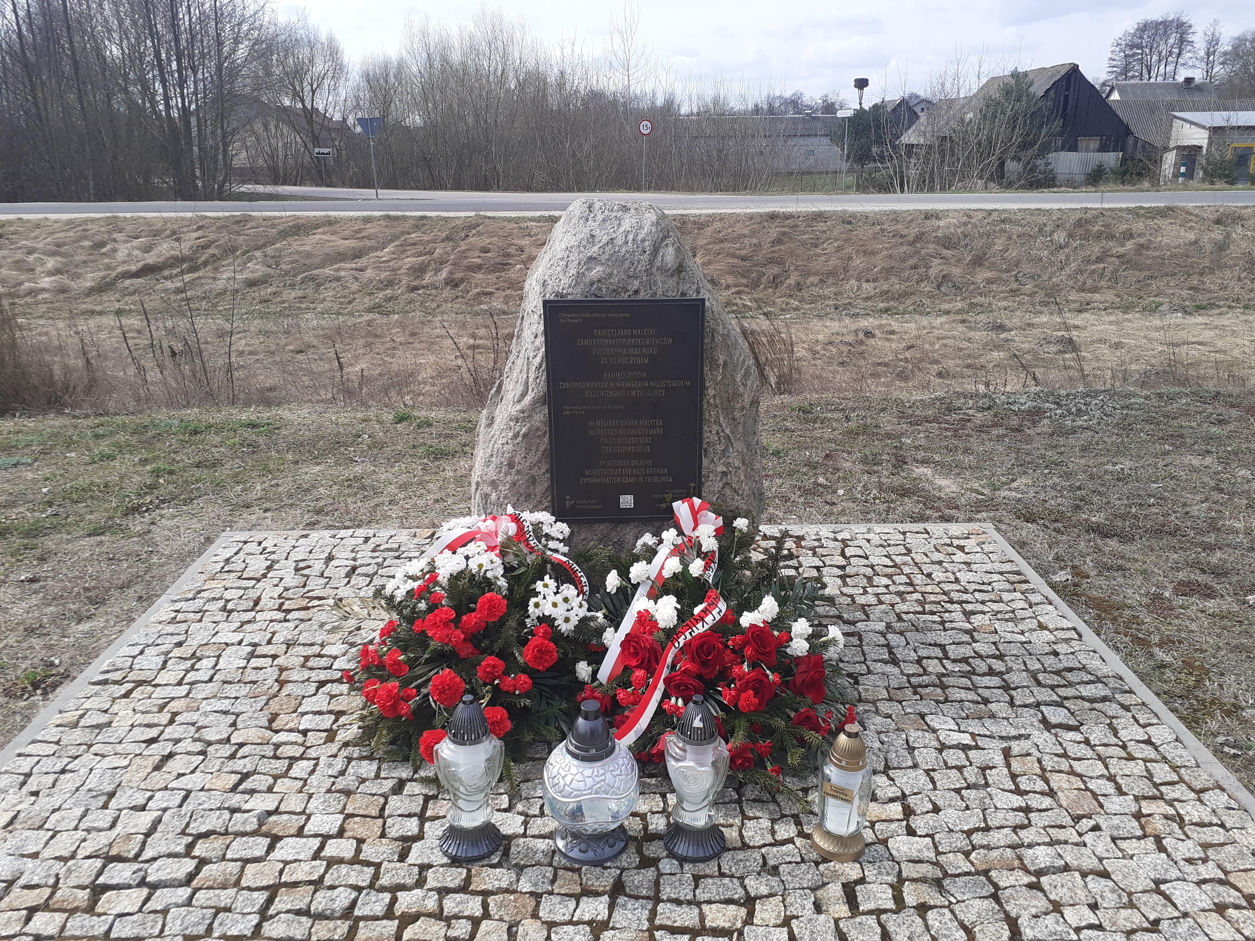 Denkmal an dem Ort der ehemaligen Bahnhofsstation im Dorf Treblinka, 31. März 2026. Die Kränze wurden vom Pilecki Institut (Witold Pilecki Institute of Solidarity and Courage) und vom Bürgermeister und den Bewohnern der Gemeinde Małkinia gestiftet; rechts eine goldene Kerze mit der goldenen Inschrift „Treblinka Museum – wir erinnern” 